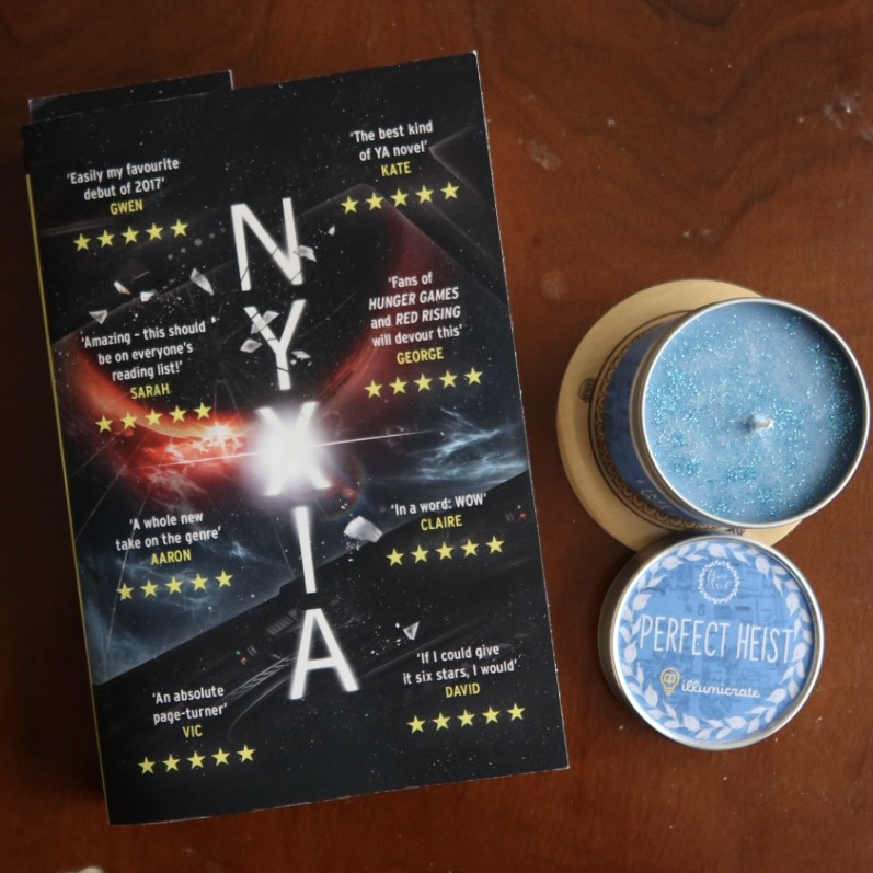 A photo of the ARC edition of Nyxia by Scott Reintgen next to a light blue and glittery Illumicrate candle